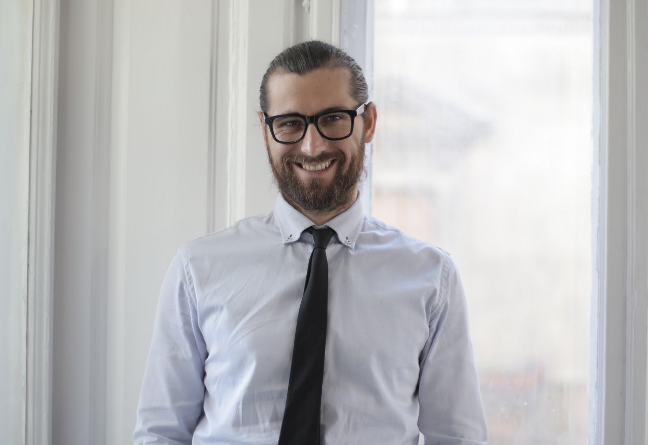 our-story Portrait of a smiling man with eyeglasses and tie, showcasing modern professional style indoors.